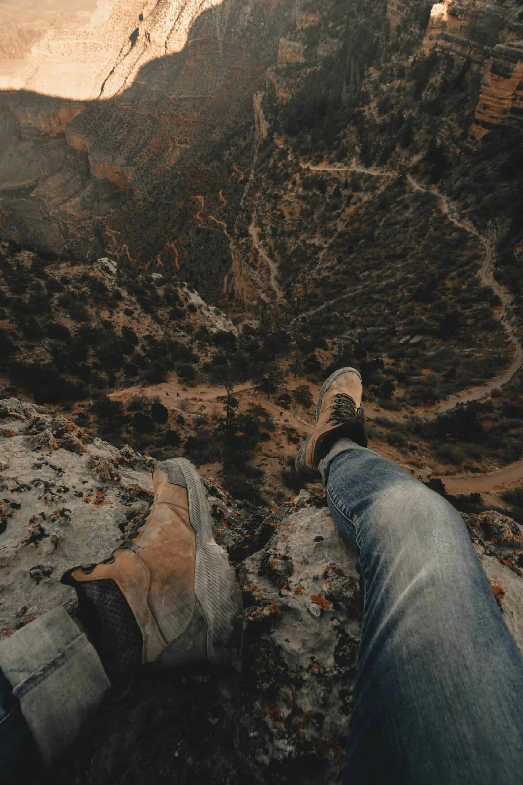 clay-banks-GyCMerrell HHiking boots worn by man sitting on edge of cliff overlooking valley below-unsplash-scaled