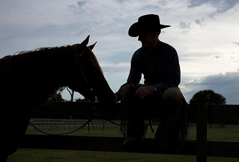 Lyle Lovett in shadow sitting on fence with his horse 
